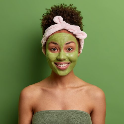 Complexion care and beauty concept. Positive woman with combed curly hair, headband, applies green moisturizing mask on face, has morning skin care routine after taking shower, isolated on green wall