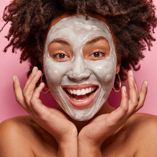 Headshot of cheerful girl with white clay mask, touches face, takes care of skin and beauty, has positive smile, Afro haircut, models over pink background, poses indoor. Facial treatment concept
